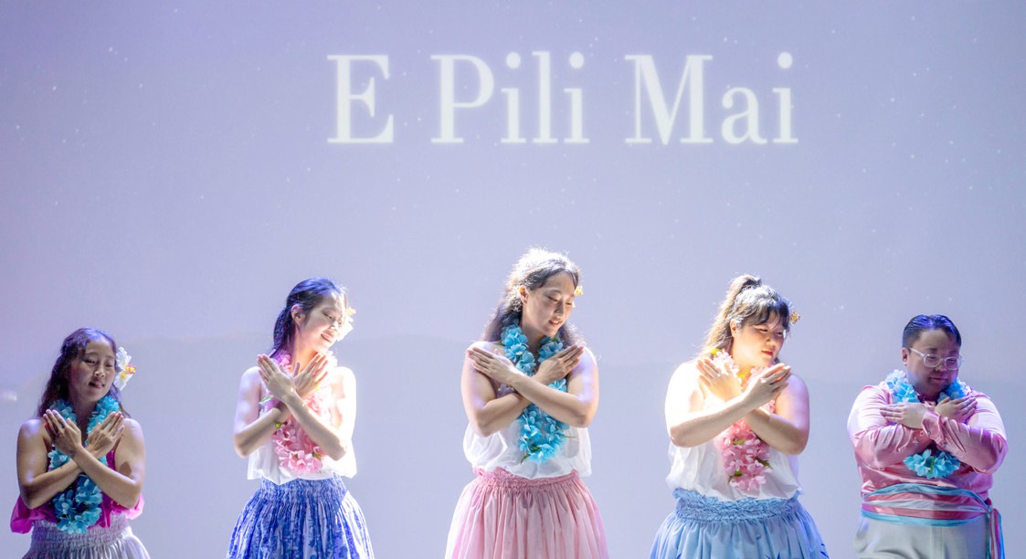 화면에 'E Pili Mai'가 띄워진 무대 앞에서 다섯 명의 멤버가 훌라춤을 추는 모습