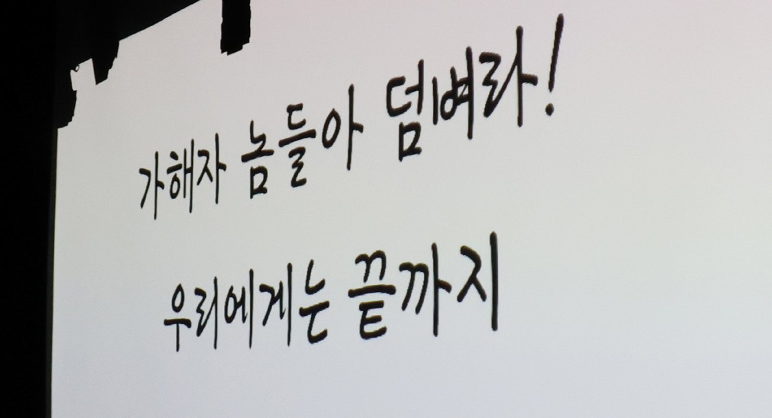 흰 스크린에 검은 손글씨로 '가해자 놈들아 덤벼라! 우리에게는 끝까지'라고 쓰인 무대 영상